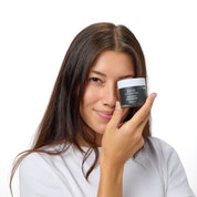 A woman with long dark brown hair holds a black cosmetic jar to her eye, wearing a white t-shirt against a solid white background.