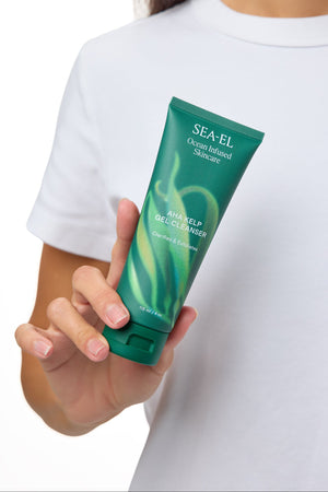 A person in a white t-shirt holds a green tube of SEA-EL Advanced Hair Regrowth Shampoo against a white background.