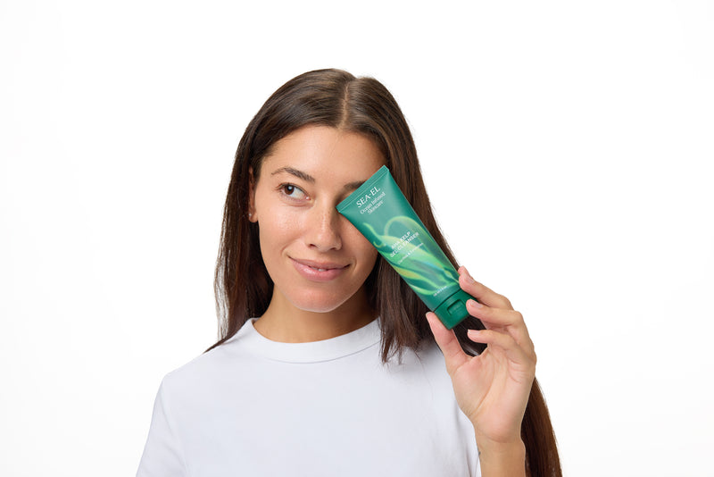 A woman with long brown hair wearing a white t-shirt holds a green tube of hair product against her eye, set against a solid white background.