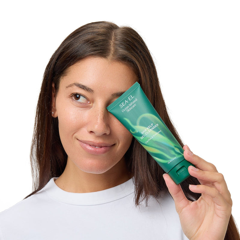 A woman with long brown hair wearing a white t-shirt holds a green tube of hair product against her eye, set against a solid white background.