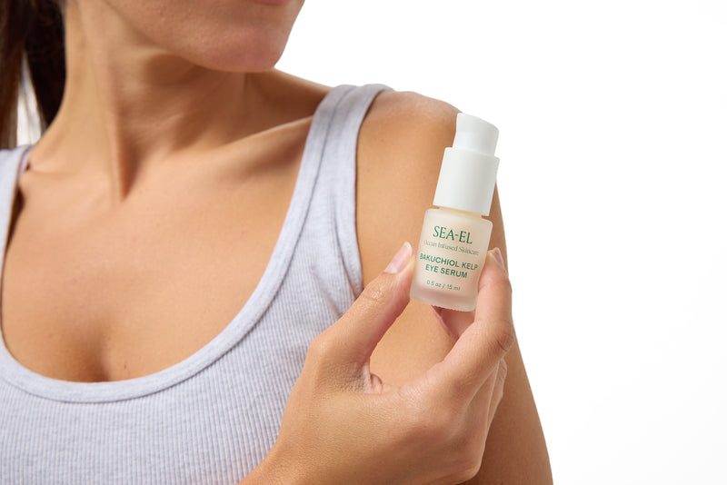 A close-up of a woman holding a bottle of SEA-EL Hagemol Kela Eye Serum against a white background.