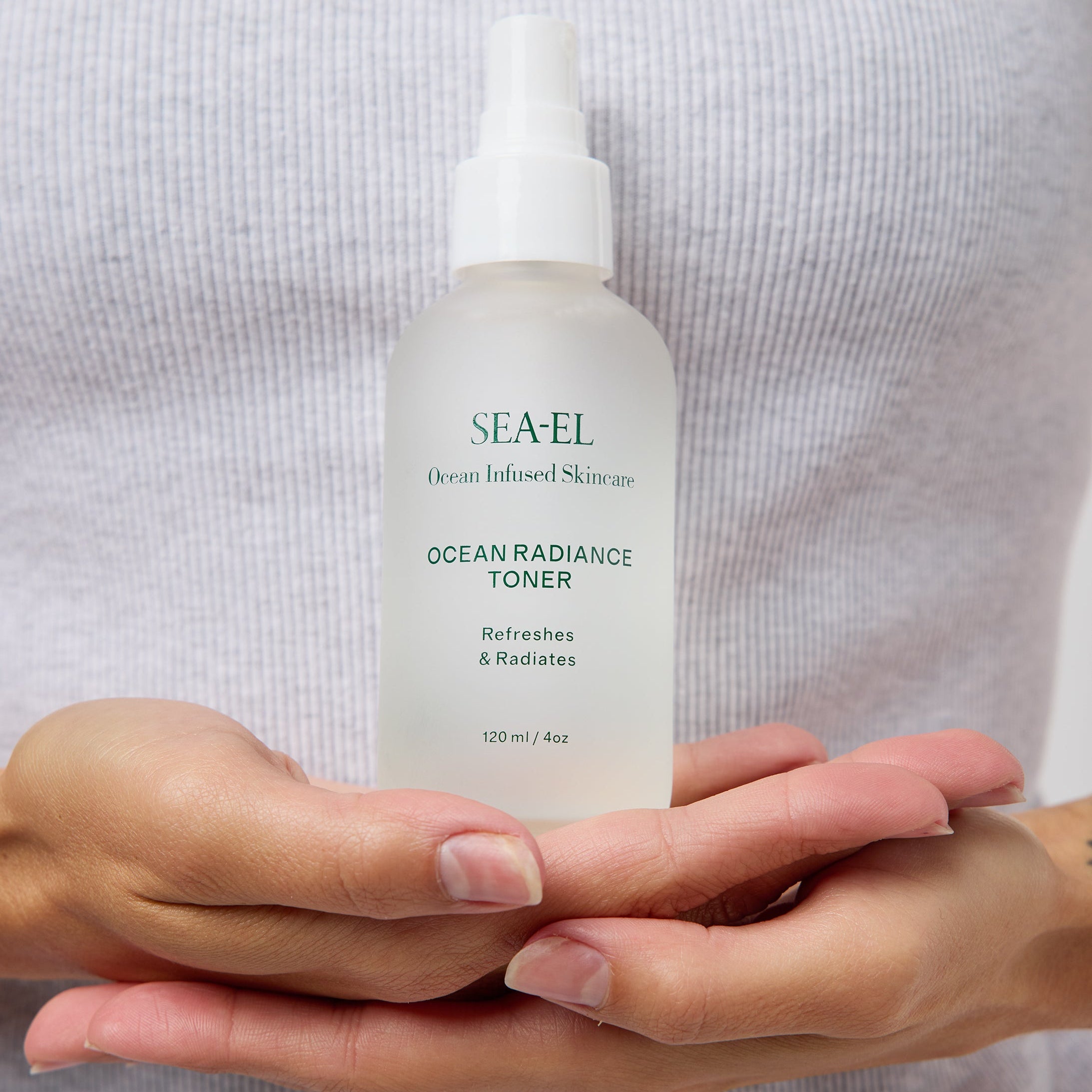 A person in a light gray ribbed top holds a frosted glass spray bottle of SEA-EL Ocean Radiance Toner. The person's hands are visible holding the bottle, and the background is a soft, out-of-focus neutral gray.