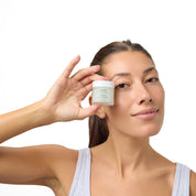 A woman holding a small white cosmetic jar near her face against a clean white background.