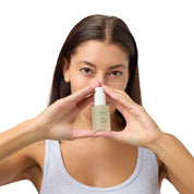 A woman with long brown hair holds a small beige cosmetic bottle in front of her face, forming a triangle with her hands. She is wearing a light gray tank top against a solid white background.