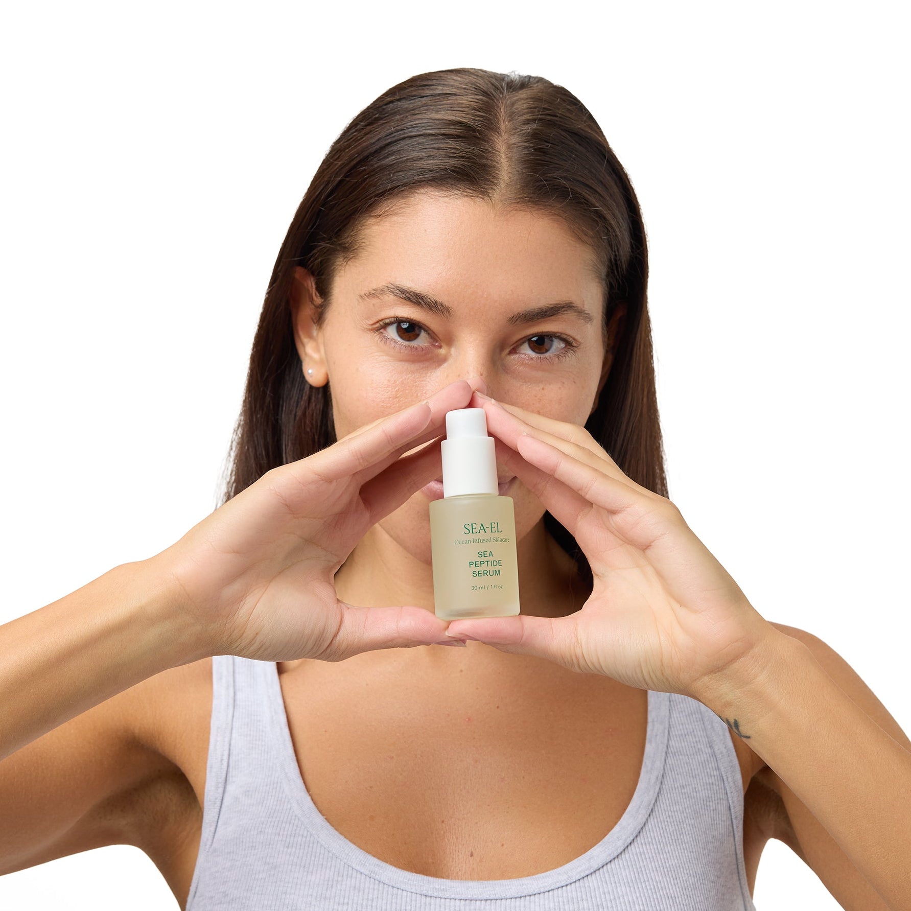 A woman with long brown hair holds a small beige cosmetic bottle in front of her face, forming a triangle with her hands. She is wearing a light gray tank top against a solid white background.
