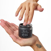 A person's hands holding and touching a small black jar of SEA-EL Sea Kelp Mud Mask against a clean white background.