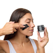 A woman with dark hair applies a black face mask to her cheek while holding a small black cosmetic jar labeled 'KAVU' next to her. She is wearing a light gray tank top against a solid white background.