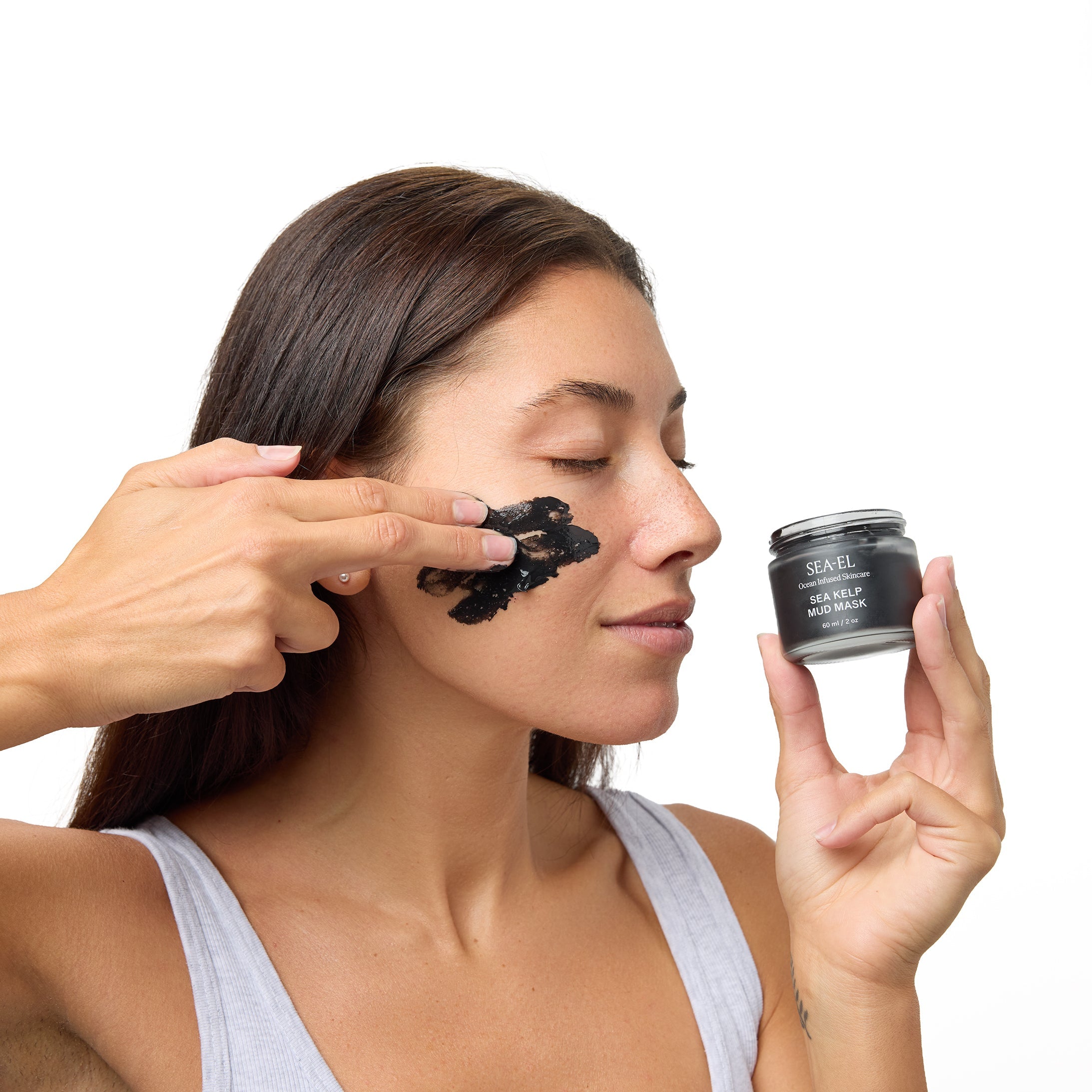 A woman with dark hair applies a black face mask to her cheek while holding a small black cosmetic jar labeled 'KAVU' next to her. She is wearing a light gray tank top against a solid white background.