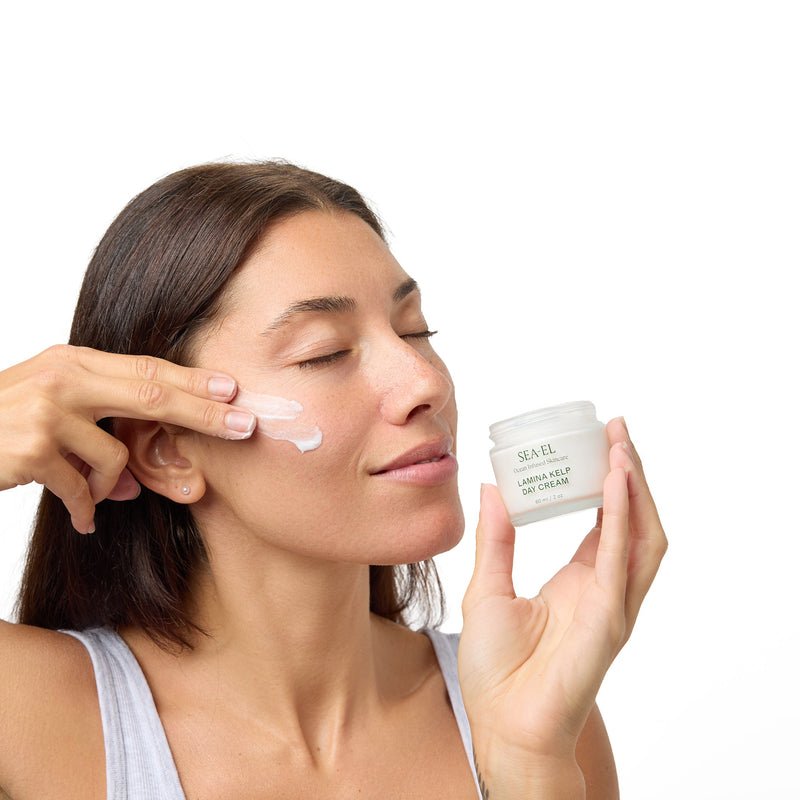 Women holding the Lamina Kelp Day Cream jar, while applying it to her cheek.
