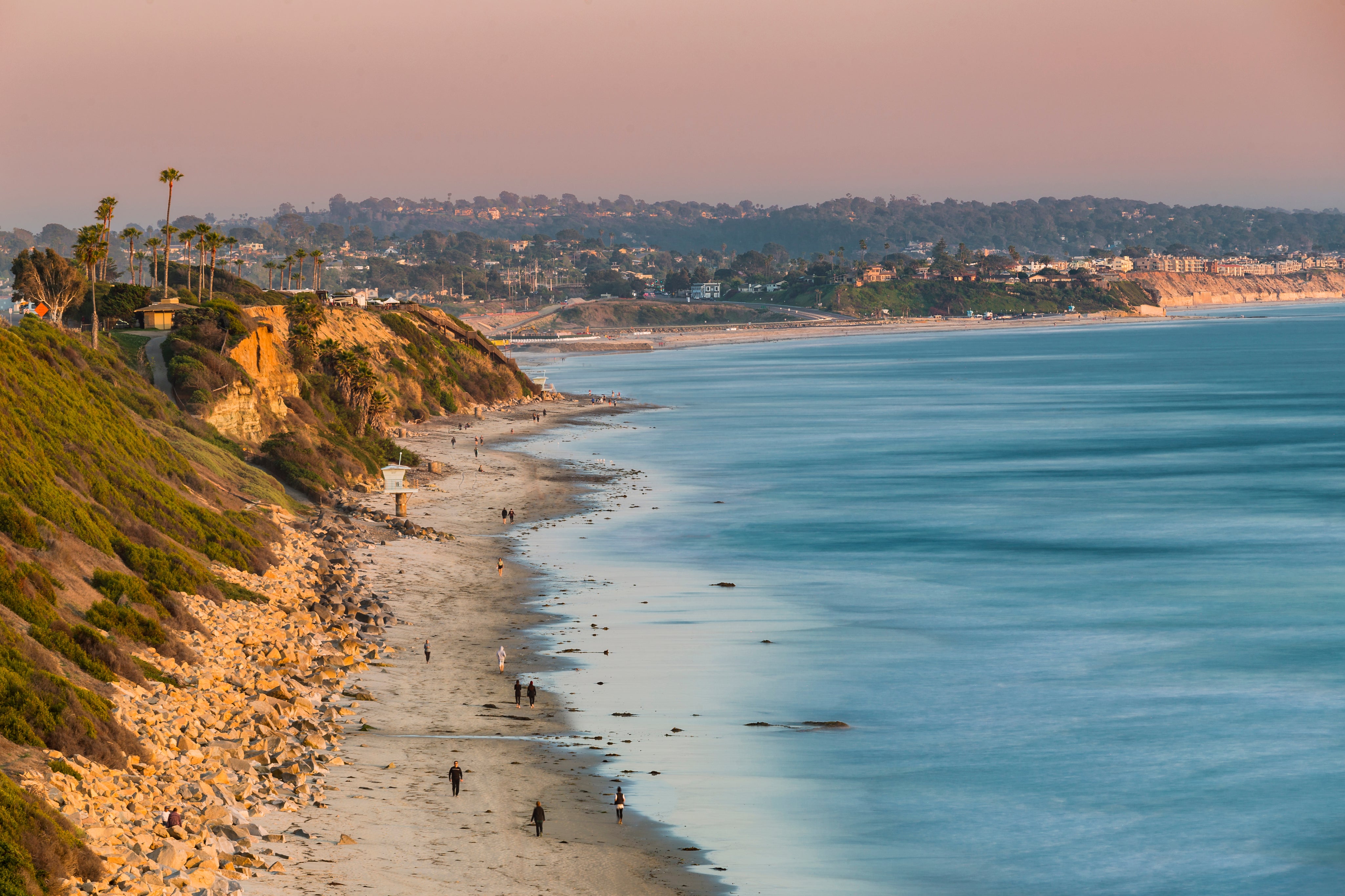 A scenic coastal landscape with a sandy beach curving along a grassy cliffside under a soft pink sky, with blue ocean water in the foreground and distant hills with houses.