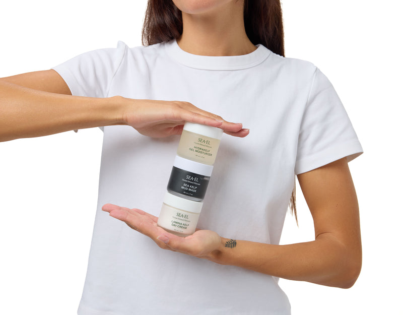 A woman wearing a plain white t-shirt holds a stack of three SAEJEL hair color bottles against a solid white background.