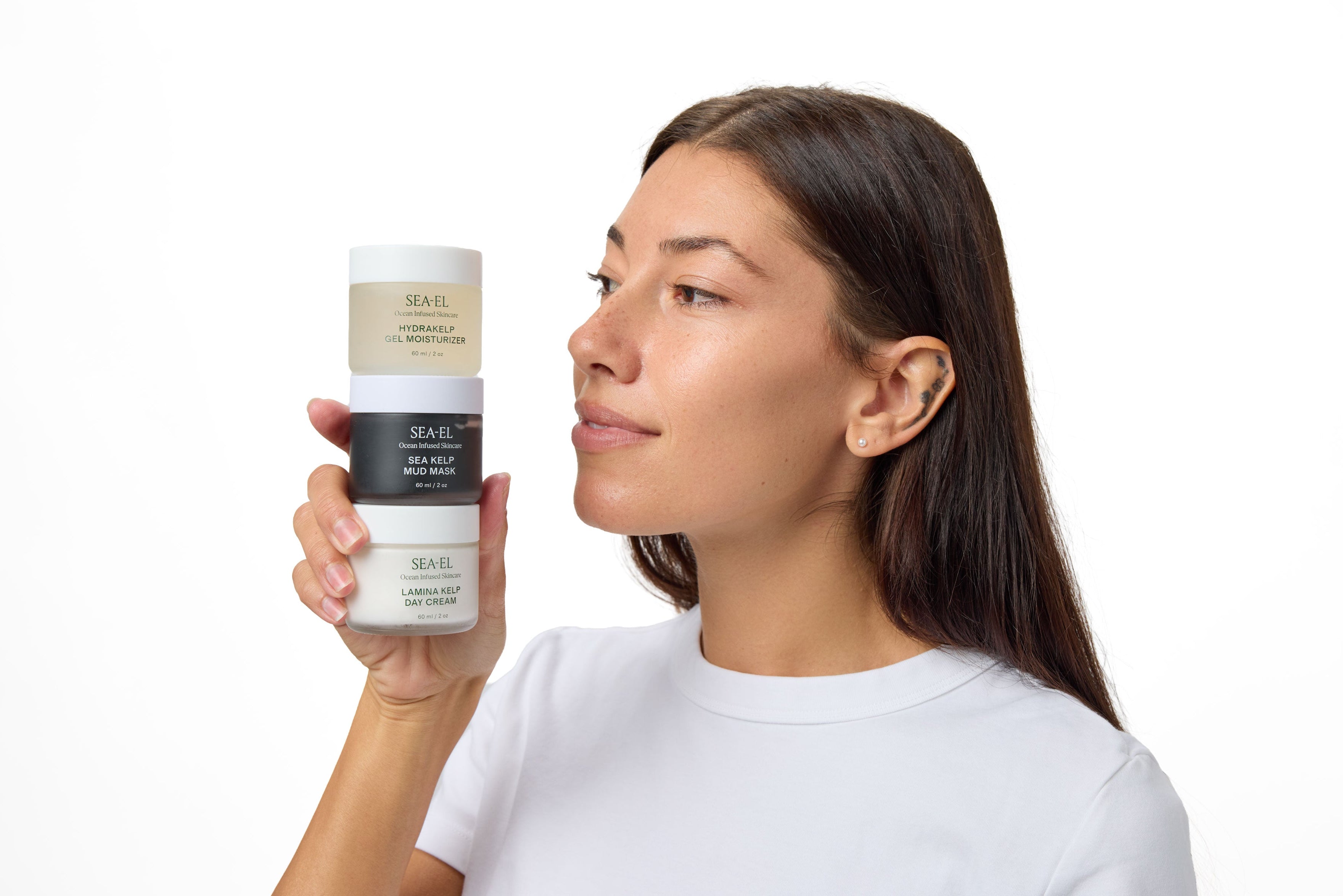 A woman with long dark hair holds a stack of three hair product jars against a plain white background. The jars are from the brand NAIL GEL, with labels in beige, black, and white.