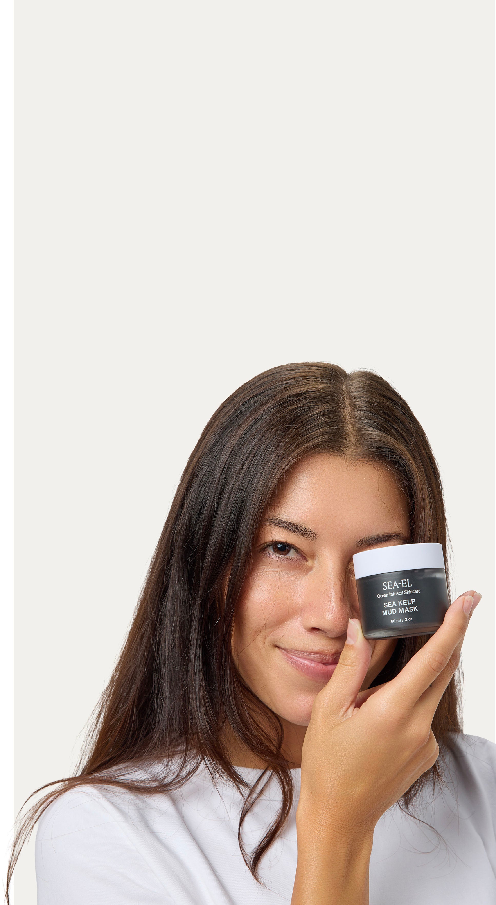 A woman with long dark brown hair holds a small black cosmetic jar near her eye against a plain light background.